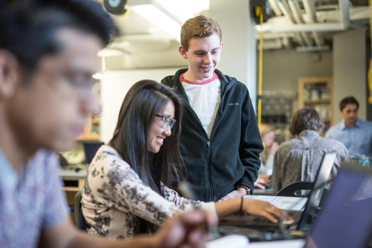 Two students collaborate on a laptop in a bustling classroom while a third student looks on from the left.