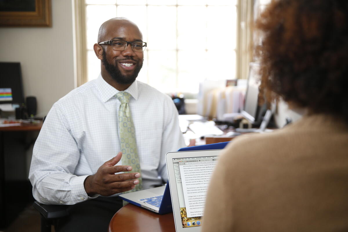 A professional black man in glasses and a tie converses with a colleague across a desk in an office setting.