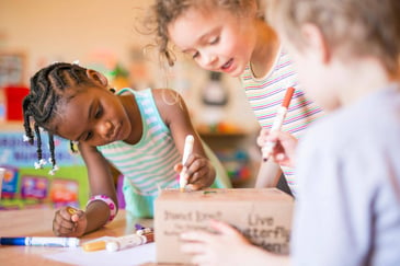 Three children are drawing on a cardboard box with markers in a classroom setting.