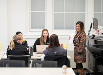 A woman stands and speaks to four seated people in a classroom setting. There are laptops and notebooks on the table.