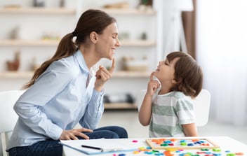 A woman and a child sit at a table facing each other, both making a shushing gesture with their fingers on their lips. The table has colorful items scattered on it.