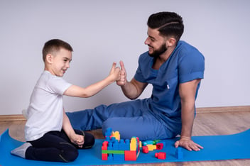 A child and a man in blue scrubs sit on a mat, smiling and high-fiving. Colorful blocks are spread on the mat.