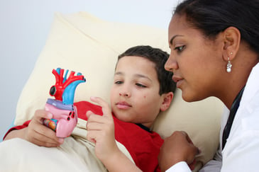 A child in bed examines an anatomical heart model while a woman, likely a healthcare professional, looks on attentively.