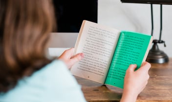 A person sitting at a desk reads a book with text visible on its pages.