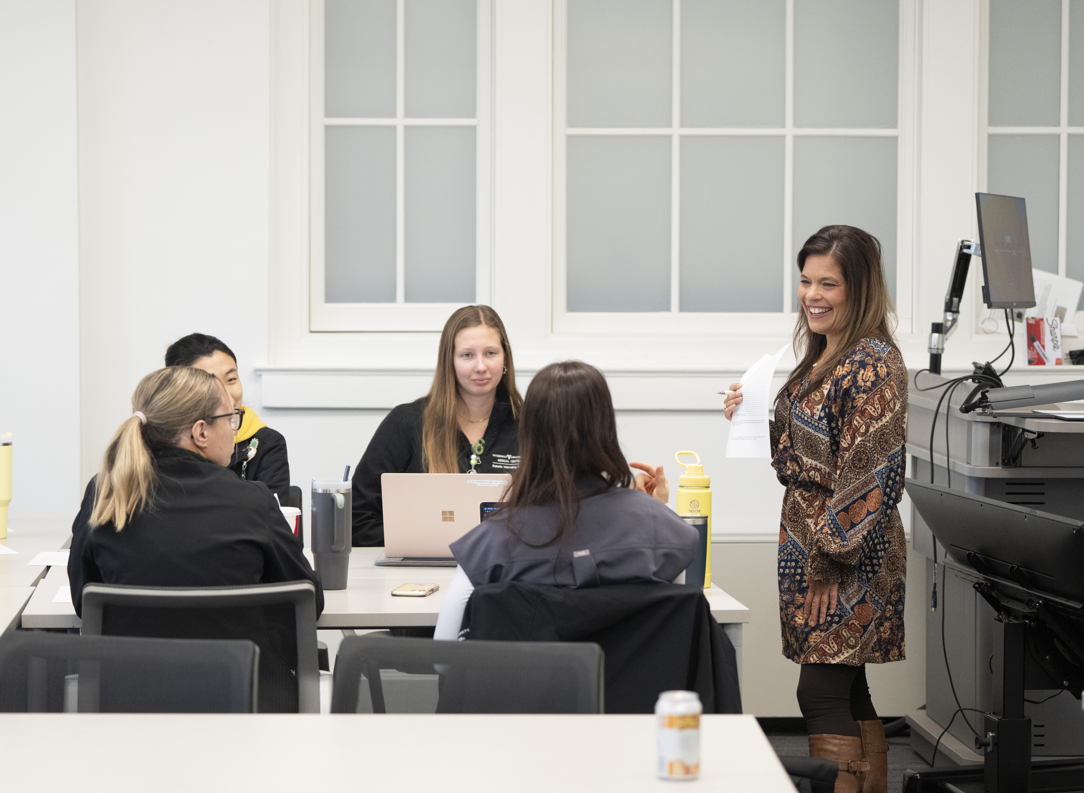 A woman stands and speaks to four seated people in a classroom setting. There are laptops and notebooks on the table.