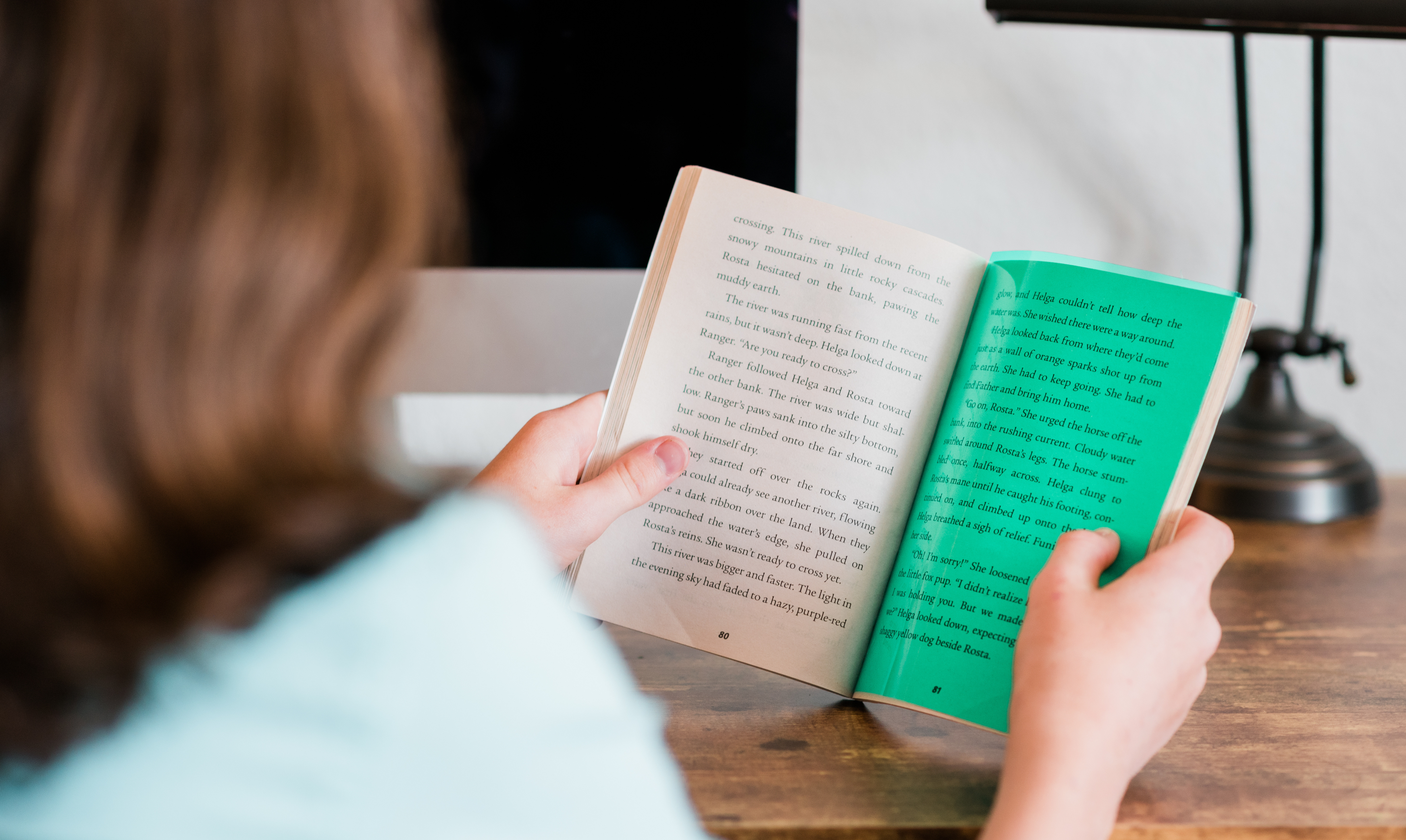 A person sitting at a desk reads a book with text visible on its pages.